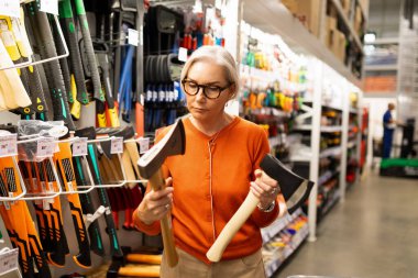 Olgun bir kadın büyük bir inşaat hipermarketinde alışveriş yaparken iki baltayı dikkatlice değerlendirir. Kaliteyi değerlendirir ve çeşitli araçlar ve ev eşyaları arasında bilgilendirilmiş seçimler yapar..