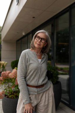 Kendine güvenen yaşlı bir kadın, gözlüklü ve açık gri hırkalı çağdaş bir binanın dışında duruyor. Gri saçları var ve çantasını tutarken nazikçe gülümsüyor..