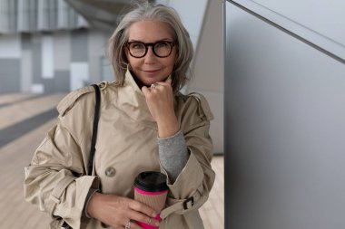 Çağdaş bir binanın yanında elinde kahve fincanı tutan şık bir kadın. Gri saçları ve gözlükleri, günün tadını çıkarırken şık trençkotunu tamamlıyor..