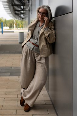Aydınlık, rahat giysiler giymiş olgun bir kadın, gün boyunca şık, kentsel bir binaya yaslanırken bir telefon görüşmesi yapıyor. Ayarlar çağdaş bir atmosferi yansıtıyor..