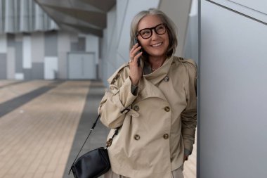 Şık bej ceketli neşeli bir kadın çağdaş bir yapıya yaslanırken bir telefon görüşmesi yapıyor. Kısa gri saçları ve gözlükleri elbisesini tamamlıyor..