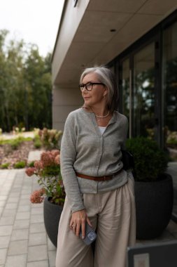 Şık, yaşlı bir kadın, yeşillik ve çiçek yataklarıyla çevrili çağdaş bir binanın dışında güneşli bir öğleden sonranın tadını çıkarıyor. Kendine güveni sofistike moda seçimini vurguluyor..