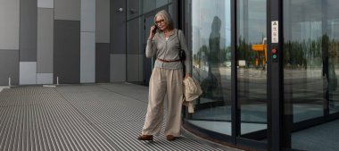 Çağdaş bir binanın döner kapısının yanında duran bir kadın sıradan bir kıyafet giyerken telefonla konuşuyor. Güneş cam yüzeylerden yansıyor..