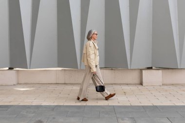 Yaşlı bir kadın çarpan gri geometrik bir duvarın yanında asfalt kaldırım boyunca yürüyor. Bej bir elbise giyiyor ve küçük siyah bir çanta taşıyor..
