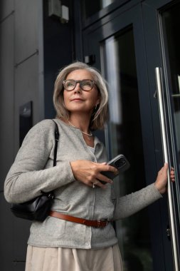 Gri saçlı ve gözlüklü bir kadın şık bir binanın girişinde duruyor. Akıllı telefonunu tutuyor ve şehre girmeye hazırlanıyor. Şehir ortamında zarafetini sergiliyor..