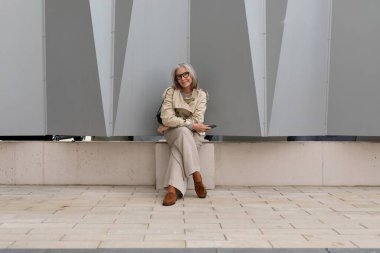 Rahat, son sınıf öğrencisi bir kadın taştan bir bankta oturmuş, açık bir günde çağdaş bir mimari yapının yanında çevresinin tadını çıkarıyor. Akıllı bir telefonu var ve mutlu bir şekilde gülümsüyor..