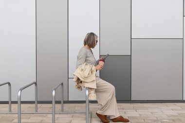 Rahat kıyafetler giymiş bir kadın, şık ve çağdaş bir binanın yanında otururken elinde içkiyle korkuluklarda rahatlar. Şehir çevresi parlak ve davetkar..