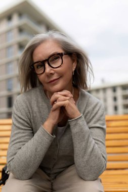 Son sınıf öğrencisi bir kadın, şehir parkındaki ahşap bir bankta oturarak eğleniyor. Gri bir hırka giymiş, kameraya sıcak bir gülümsemeyle bakıyor, gümüş saçlarını ve gözlüklerini gösteriyor..