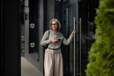 Gri saçlı ve gözlüklü bir kadın şehir ortamında şık ve çağdaş bir binanın önünde duruyor. Kapının önünde telefonuna bakıyor, kendine güveni tam..