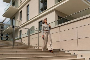 Aydınlık giyinmiş bir kadın güneşli bir öğleden sonra çağdaş bir binanın merdivenlerinden aşağı iniyor. Akıllı bir telefon taşıyor ve kendine güveni ve tarzı var..