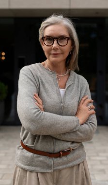 Kendinden emin yaşlı bir kadın kollarını kavuşturarak çağdaş bir binanın önünde duruyor. Gri saçları yüzünü çerçeveler, gözlük ve şık kıyafetlerle tamamlanır. Çiçek ekiciler ortama zarafet katar..