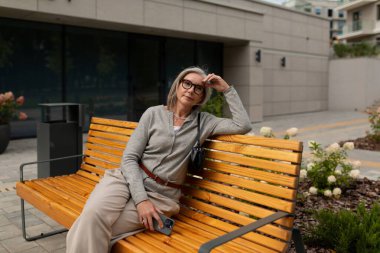 Gözlüklü orta yaşlı bir kadın, yeşil ve modern mimari ile dolu bir kentsel alanda rahatlamak için, parlak ahşap bir bankta gelişigüzel oturuyor..