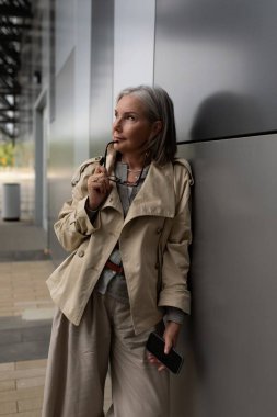 Gri saçlı şık bir kadın, çağdaş bir şehir bölgesinde parlak bir duvara yaslanıyor, telefonunu tutuyor ve derin düşüncelere dalmış, açık, zarif giysiler giymiş..