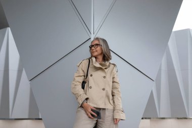 Gri saçlı bir kadın gri bir duvara karşı poz veriyor, bej renkli trençkot giyiyor ve şık gözlükler takıyor. Bir telefon tutuyor, şehir ortamında güven ve modernlik yayıyor..