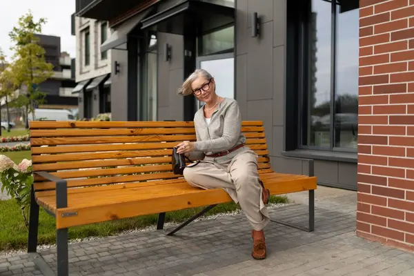 Kısa gri saçlı neşeli yaşlı bir kadın modern bir binanın dışındaki ahşap bir bankta rahatça oturuyor. Küçük bir torba tutarken gülümsüyor, sakin atmosferin tadını çıkarıyor..