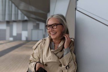 Gülümseyen bir kadın şık bir yapı karşısında duruyor, şık bir trençkot ve gözlük takıyor. Güneşli bir günde canlı bir şehir ortamında mutluluk saçar..