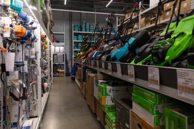 Kalabalık bir tamirhanenin koridorlarını elektrikli aletler, bahçe malzemeleri ve bahçe malzemeleri dolduruyor. Müşteriler araç ararken, çalışanlar ürünleri düzenli bir şekilde depoluyorlar..
