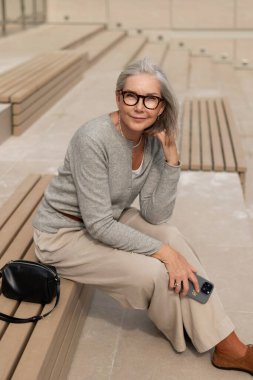 Gözlüklü yaşlı bir kadın, açık hava alanındaki ahşap banklarda gelişigüzel oturur. Açık renk bir süveter ve pantolon giymiş, çevresinin tadını çıkarıyor..