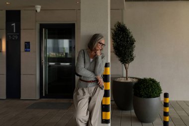 Son sınıf öğrencisi bir kadın, çağdaş bir binanın dışındaki sarı çizgili bir direğin yanında durup gündüz vaktinin tadını çıkartıyor. Rahat ve düşünceli görünüyor..