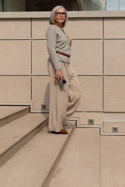Kendine güvenen bir kadın düz malzemelerden yapılmış açık renkli basamakların tepesinde duruyor. Rahat bir süveter ve şık bir pantolon giyer, şehir manzarasında güneşli bir günün tadını çıkarır..