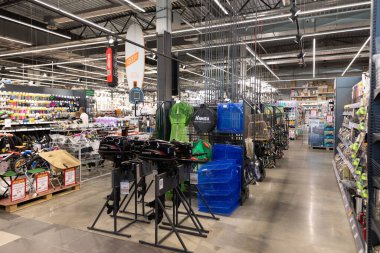 Geniş bir hırdavatçıda raflar çeşitli aletlerle, temizlik malzemeleriyle ve tamir malzemeleriyle doludur. Müşteriler mağaza saatlerinde ev geliştirme çözümleri aramak için koridorları tarıyorlar.