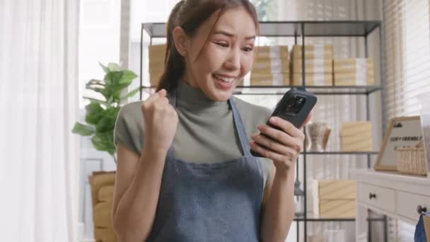 中小企業は緑のローンお金がネットゼロ小売店のために承認行く Asia Gen Zの人々若い大人の女性スマートエコフレンドリーな小さな店の所有者は良いニュースの結果を読む家のオフィスで携帯電話を見て幸せな売り手 — ストック動画