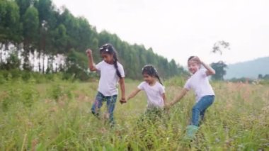 Three active sister cute child girls hold hands move way forward go walk in rural green grass field. Happy sibling young kids group asia people relax smile joy explore fun in love care nature forest.