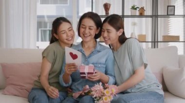 Mother's day two grown up child cuddle hug give flower gift box red heart card to mature mum. Love kiss care mom asia middle age adult three people sitting at home sofa happy smile enjoy family time.