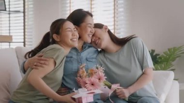 Mother day two grown up child cuddle hug give flower gift box red heart card to mature mum. Love kiss care mom asia middle age adult three people sitting at home sofa happy smile enjoy family time.