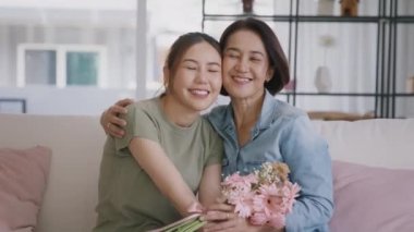 Happy time Mother day grown up child girl looking at camera cuddle hug give flower bouquet gift to mature mum. Love kiss care mom asia middle age adult people smile enjoy relax sitting at home sofa.