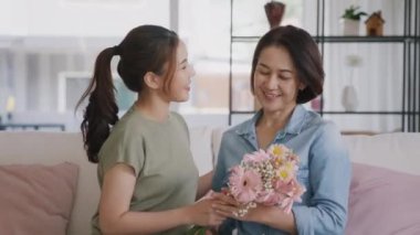 Happy time Mother day grown up child girl looking at camera cuddle hug give flower bouquet gift to mature mum. Love kiss care mom asia middle age adult people smile enjoy relax sitting at home sofa.