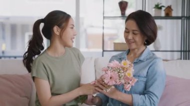Happy time Mother day grown up child girl looking at camera cuddle hug give flower bouquet gift to mature mum. Love kiss care mom asia middle age adult people smile enjoy relax sitting at home sofa.