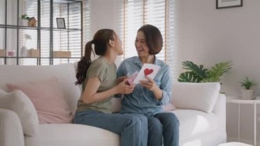 May Mother's day young adult grown up child cuddle hug give flower gift box red heart card to mature middle aged mum. Love kiss care mom asia people sitting at home sofa happy smile enjoy family time.