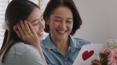 May Mother's day young adult grown up child cuddle hug give flower gift box red heart card to mature middle aged mum. Love kiss care mom asia people sitting at home sofa happy smile enjoy family time.