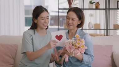 May Mother's day young adult grown up child cuddle hug give flower gift box red heart card to mature middle aged mum. Love kiss care mom asia people sitting at home sofa happy smile enjoy family time.