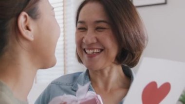 May Mother's day young adult grown up child cuddle hug give flower gift box red heart card to mature middle aged mum. Love kiss care mom asia people sitting at home sofa happy smile enjoy family time.