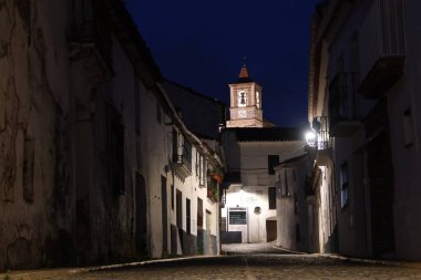 Castano del Robledo, Huelva, İspanya, 27 Mayıs 2023: Endülüs 'ün büyülü şehri Castano del Robledo' nun aydınlatılmış caddesindeki evler ve kilise çan kulesi