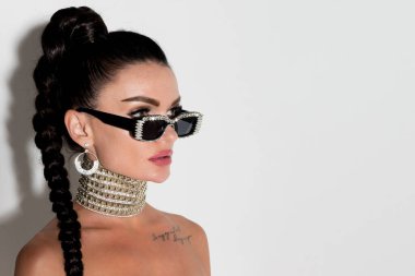 Sofistike ve tarz sahibi, güneş gözlüklü ve dalgalı at kuyruklu bir kadın yakın çekim stüdyo resminde büyüleyici bir poz veriyor, cazibesi inkar edilemez.