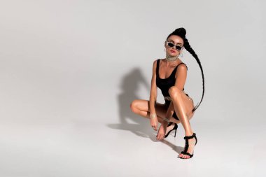 Özgüven ve cazibeyle poz veren ince bir bayan stüdyo zemininde siyah iç çamaşırlarıyla oturur gergin vücudu ve dalgalı at kuyruğu bu büyüleyici kompozisyonda dikkatleri üzerine çeker.