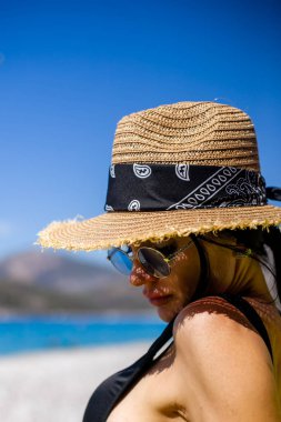 Beyaz mayo giymiş, güzel şapkalı, turkuaz deniz manzaralı beyaz kumsalda oturan genç bir kadın denize doğru bakıyor.