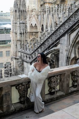 Şehir manzarası zarafeti: Güzel genç bir kadın, kentsel arkaplanı tamamlayan beyaz kürkü ile Milano Katedrali 'nin çatısında zarifçe poz veriyor.