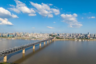 Wuhan Çin şehri Yangtze Nehri boyunca güzel bir şekilde uzanır, açık mavi gökyüzü altında beyaz bulutlarla geniş suları geçen uzun bir köprüyle.