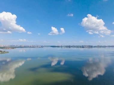 Geniş bir göl manzarası güzel bir şekilde parlak mavi gökyüzünü ve pofuduk beyaz bulutları yansıtır, açık güneşli bir günde uzak bir şehir siluetine doğru uzanır..