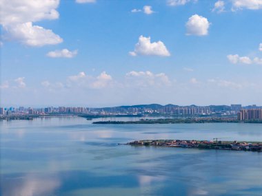 Geniş Tangxun Gölü, Jiangxia Bölgesi, Wuhan, Çin 'deki uzak şehir yapılarını ve doğal adaları gösteren beyaz bulutlu parlak mavi bir gökyüzünü yansıtır..