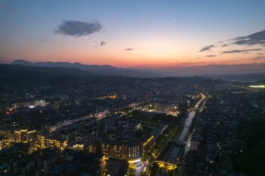 Akşam ışıkları Çin 'in Wudang kasabasını aydınlatıyor. Şehir manzarası boyunca uzanan binalar ve nehir renkli gökyüzünün altındaki uzak dağlarla aydınlanıyor..