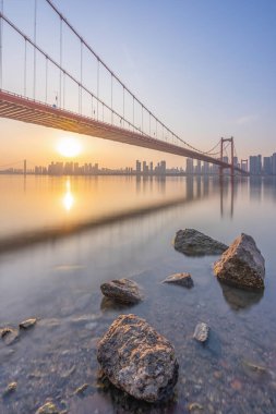 Büyük bir köprü, parlak bir gökyüzünün altında şehir silueti olan bir nehri geçer..