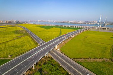 Yükseltilmiş otoyollar güzel bir şekilde kesişiyor canlı sarı çiçek tarlaları, hareketli modern şehir siluetine ve büyük bir nehir köprüsüne bağlanıyor..