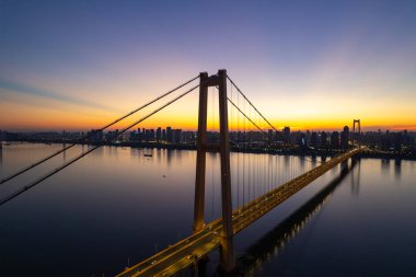 Güneş şehrin üzerinde batarken, gökyüzünü aydınlatan görkemli bir köprü geniş bir nehir boyunca uzanır..