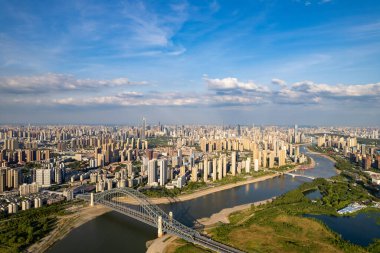 Wuhan Çin şehir manzarası parlak gökyüzü altında nehir kıyıları boyunca geniş kentsel gelişim gösteriyor. Yüksek binalar ufukta suyla dolu. Yeşil ağaçlar ön planda ve nehir adalarında. 
