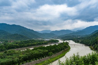 Dujiangyan nehri manzarası Sichuang Çin sarmal su yollarını gösteriyor. Etrafı yemyeşil dağlarla çevrili bulutlu bir gökyüzü altında, suyu kaplayan uzun bir yapısı var..